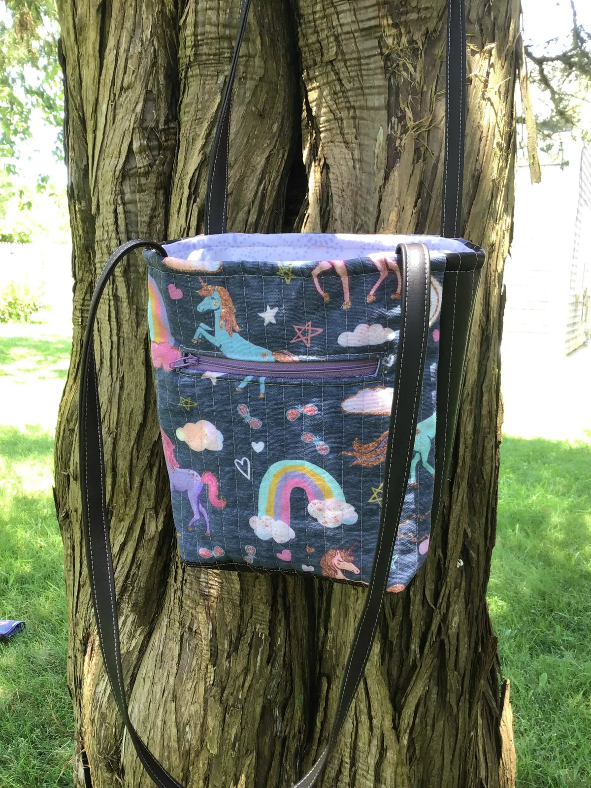 A navy unicorn print bag with a rainbow and clouds hangs on a tree.