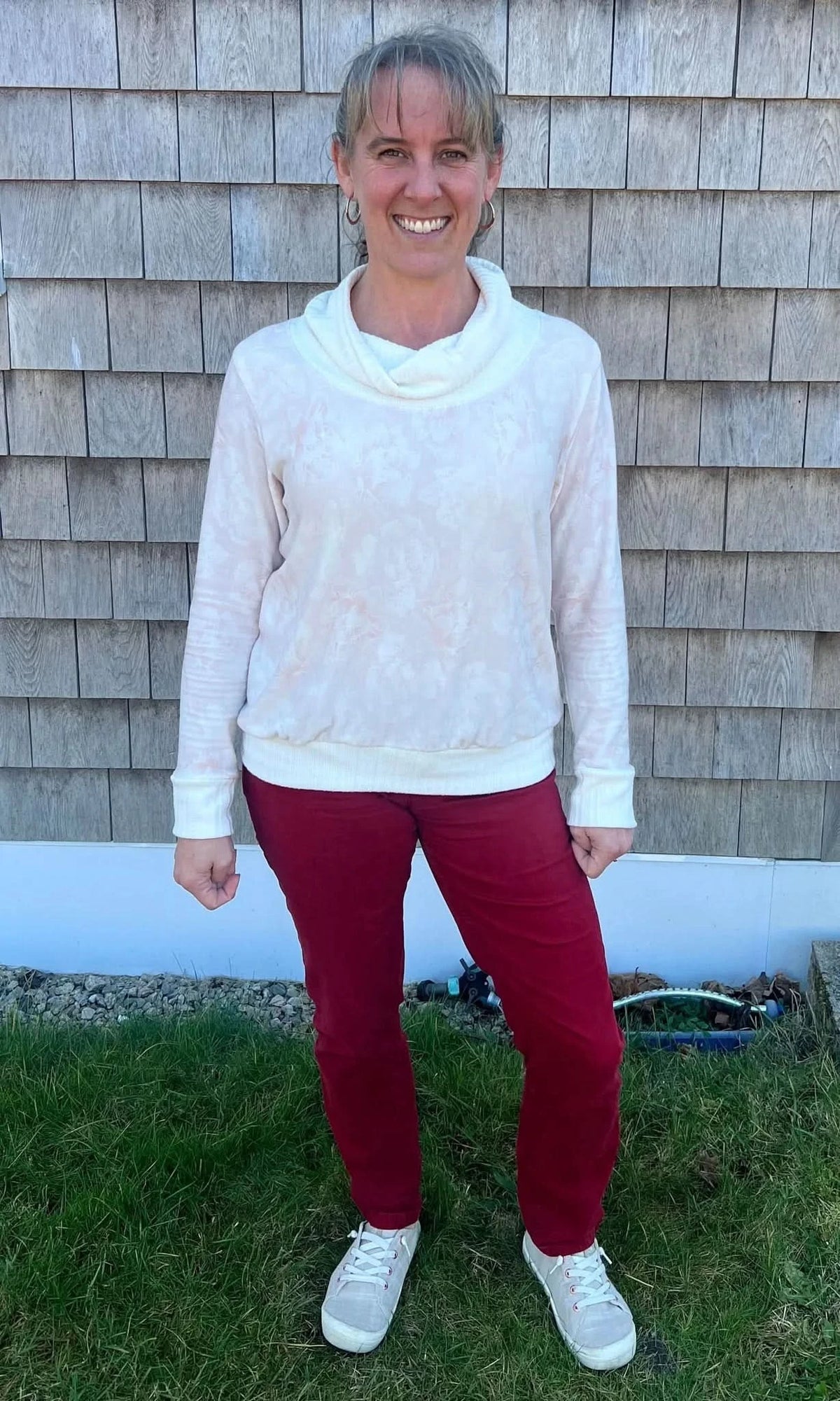 A woman smiles wearing a light pink floral cowl neck top and red pants.