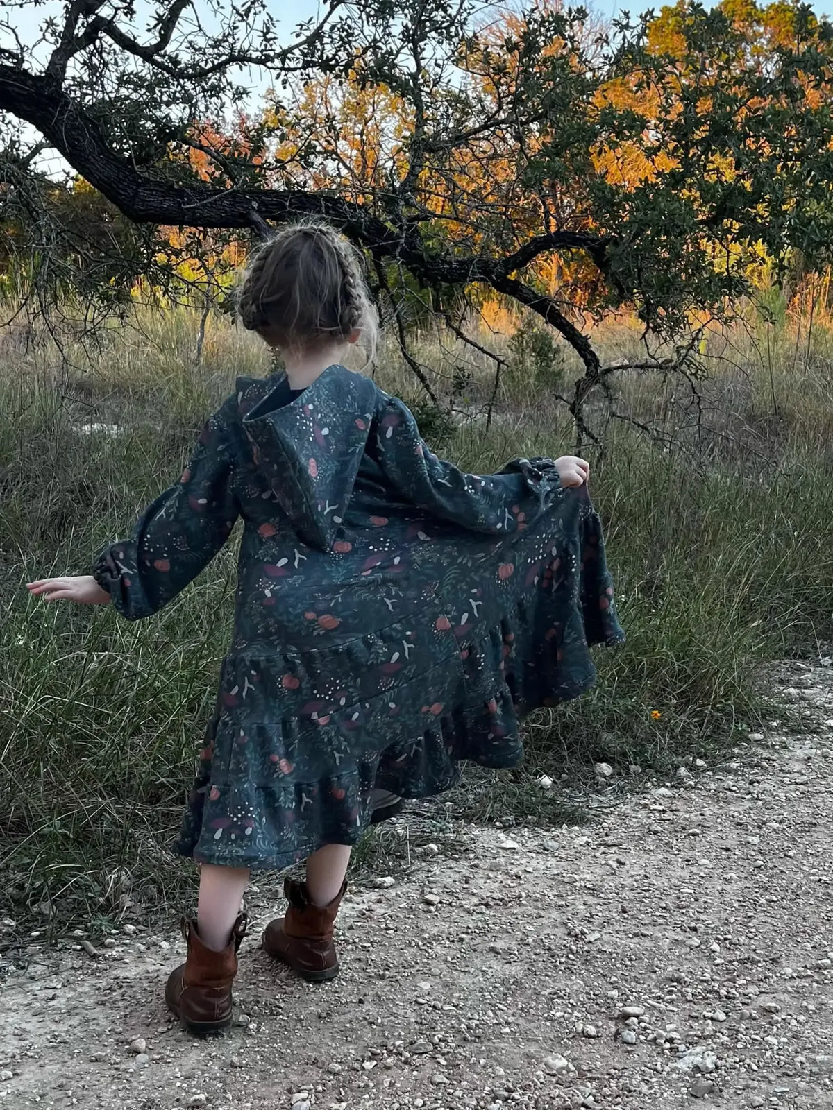 A child in a green dress with a forest foliage pattern walks on a dirt path.