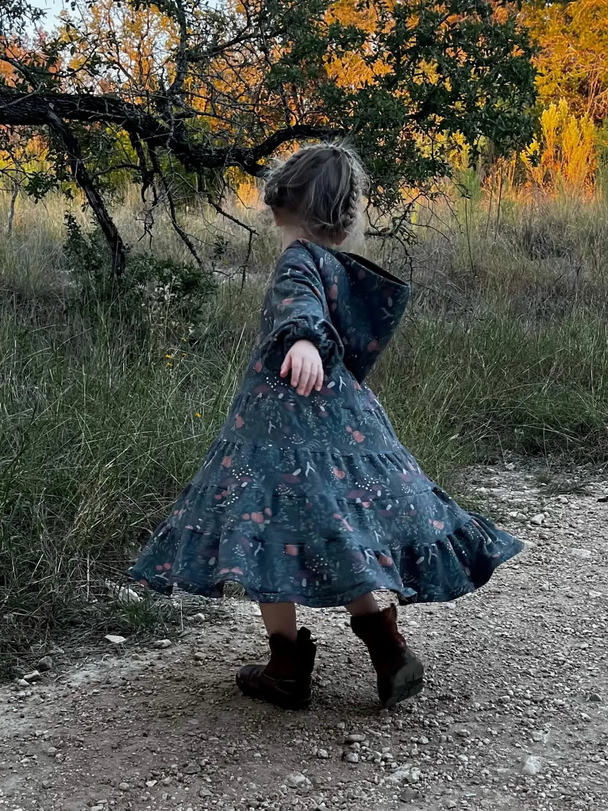 A child in a green dress with a forest foliage pattern twirls on a dirt path.