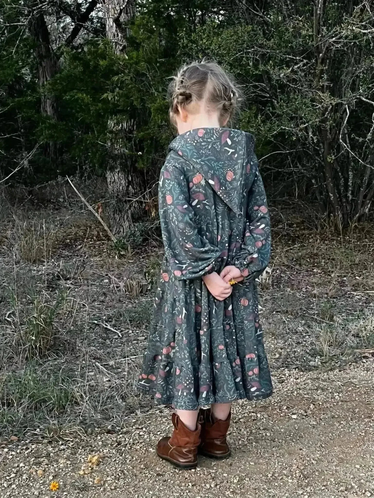 Girl in green botanical print dress with hood and brown boots in forest.