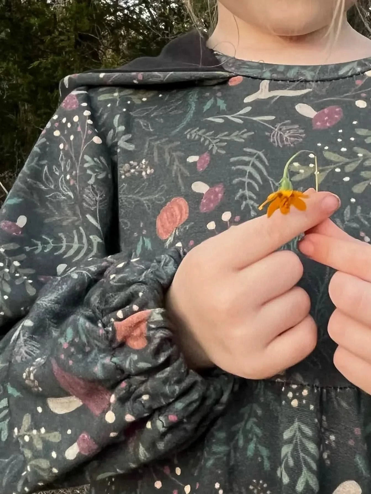 A child in a green forest foliage print dress holds a small orange flower.