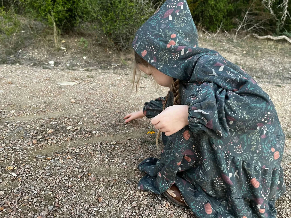 A child in a green forest foliage print dress with a hood picks a yellow flower.
