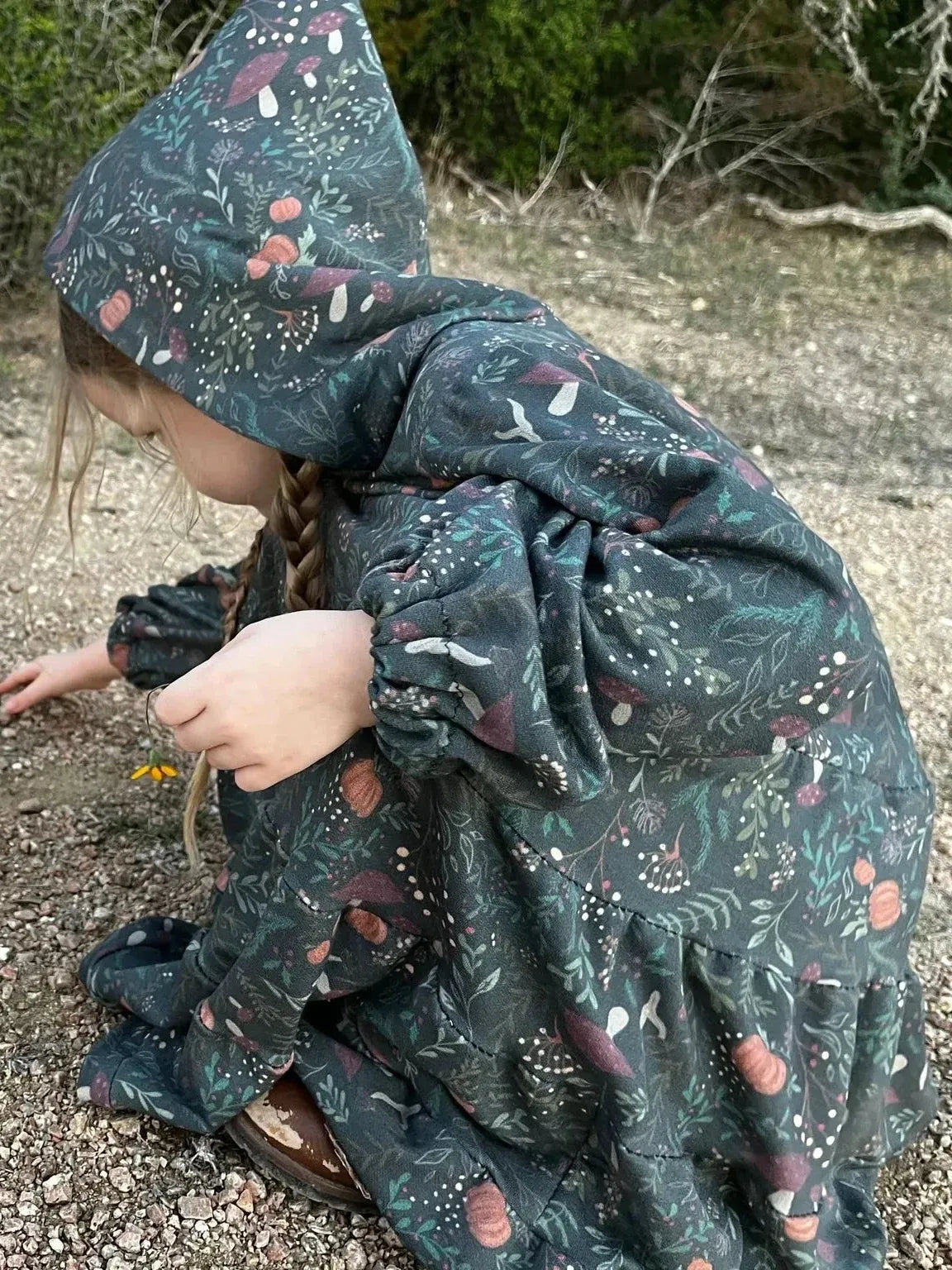 Child in a green botanical print dress with a hood, kneeling on a gravel path.