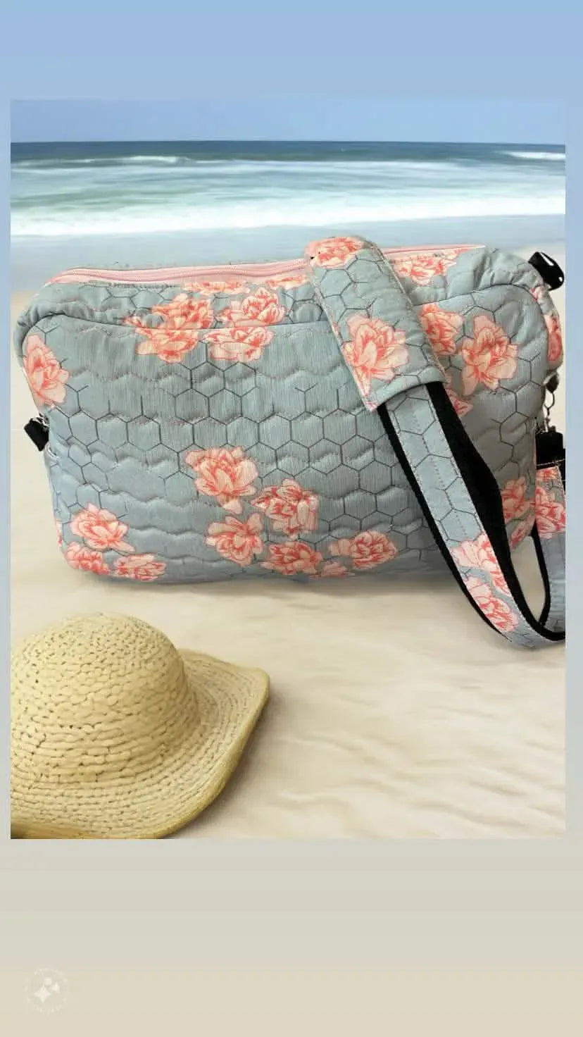 A quilted blue bag with a honeycomb pattern and pink flowers sits on a sandy beach.