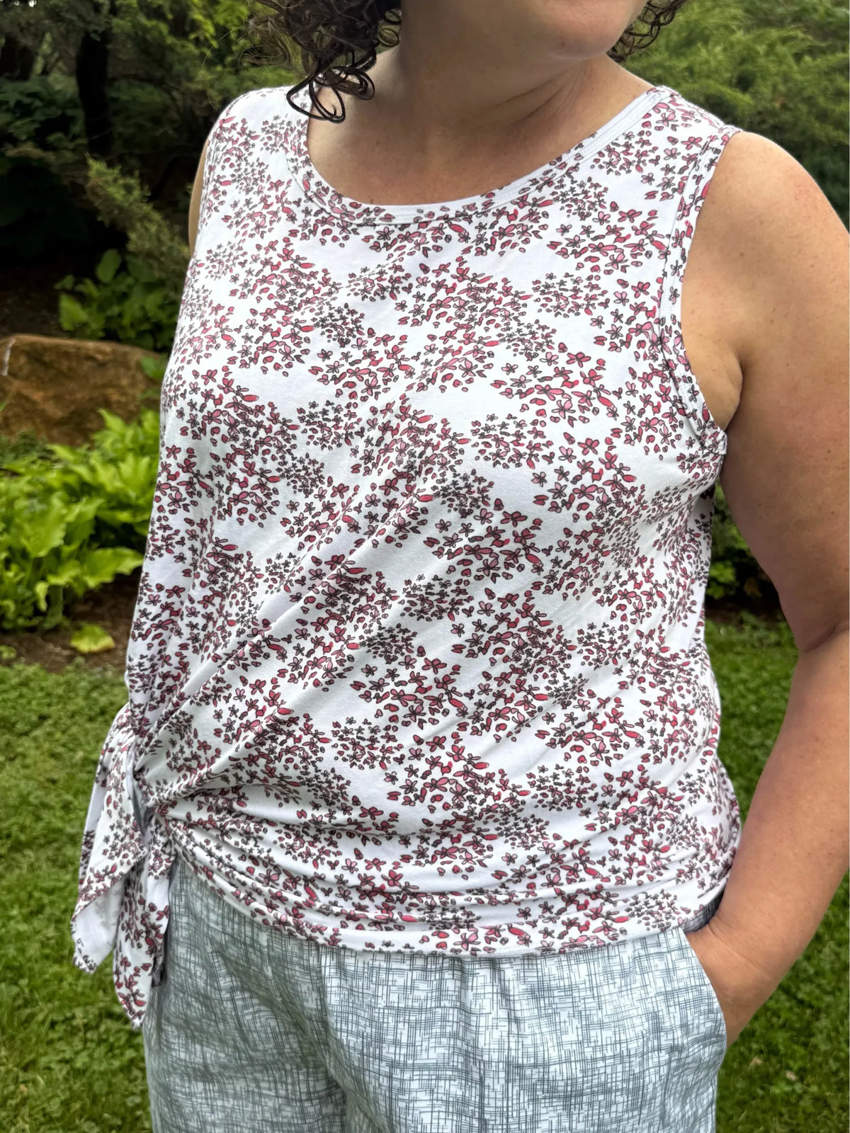A woman wears a white tank top with a red floral pattern and light gray patterned pants.