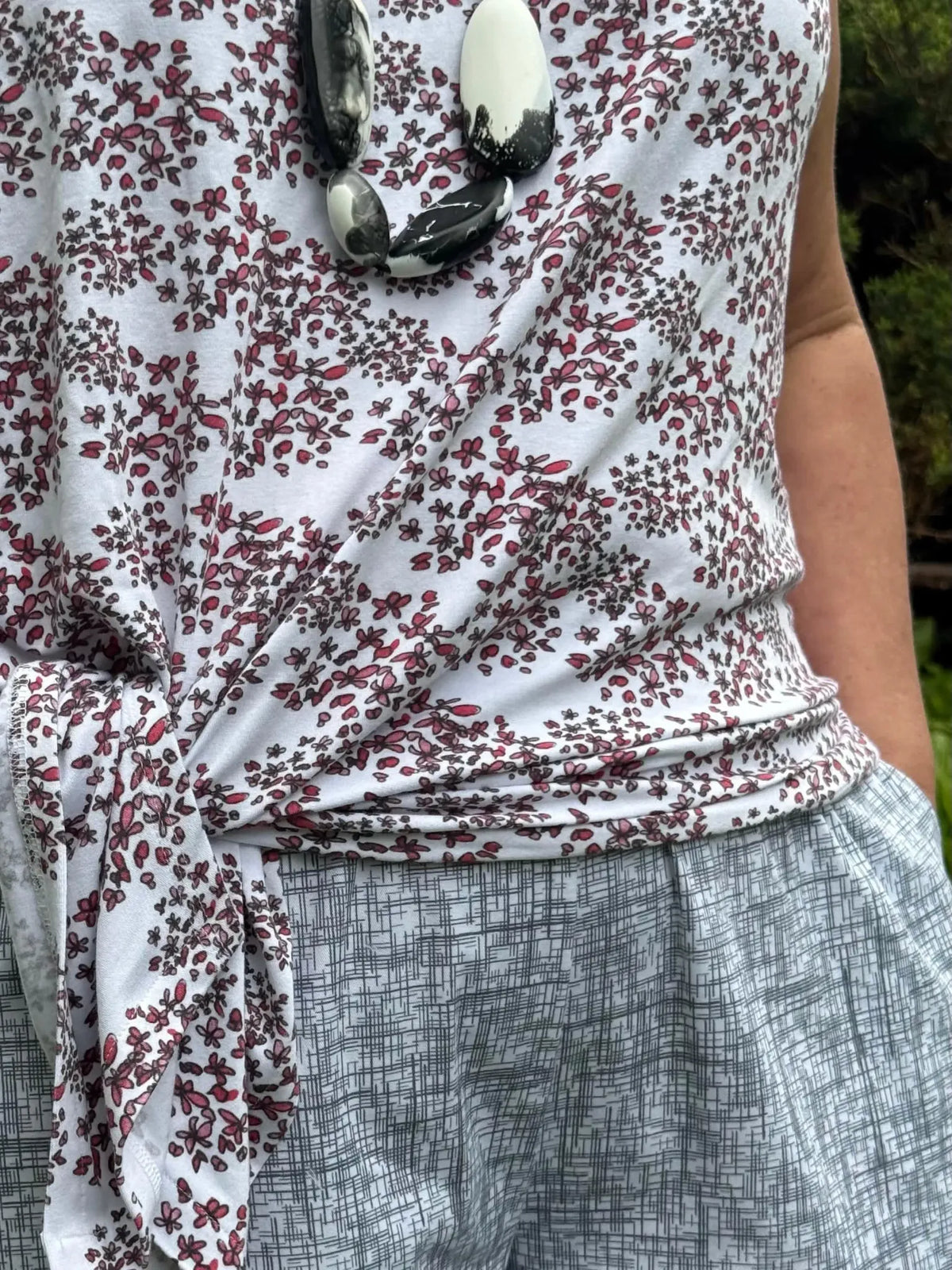 A person wears a white floral top with a black and white necklace and patterned skirt.