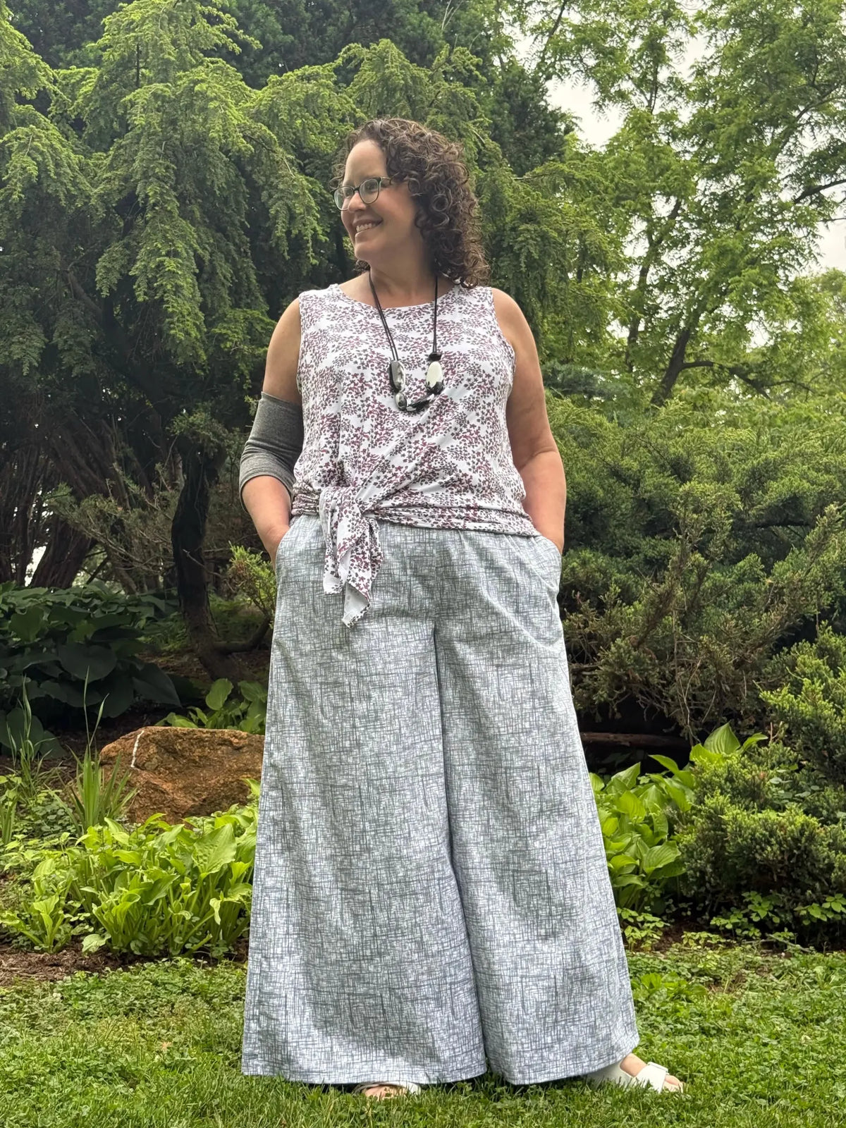 A woman in a patterned top and wide-leg pants stands in a garden.