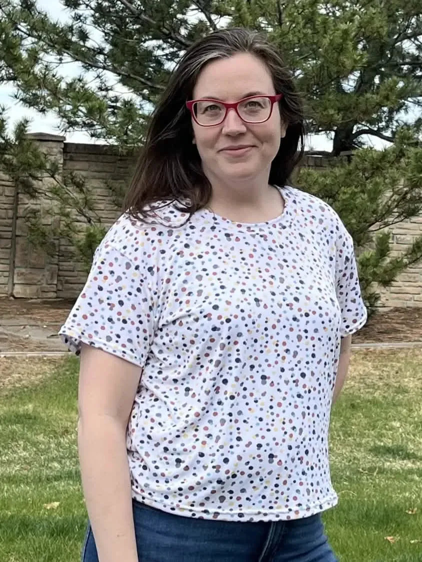 A woman wears a white t-shirt with colorful speckles, red glasses, and blue jeans.