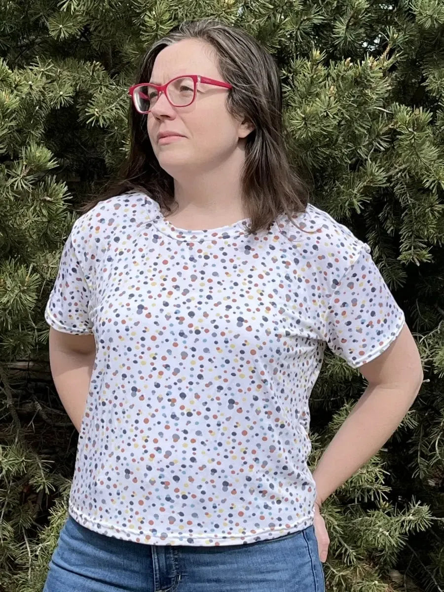 A woman wears a white shirt with colorful sprinkles, blue jeans, and red glasses.