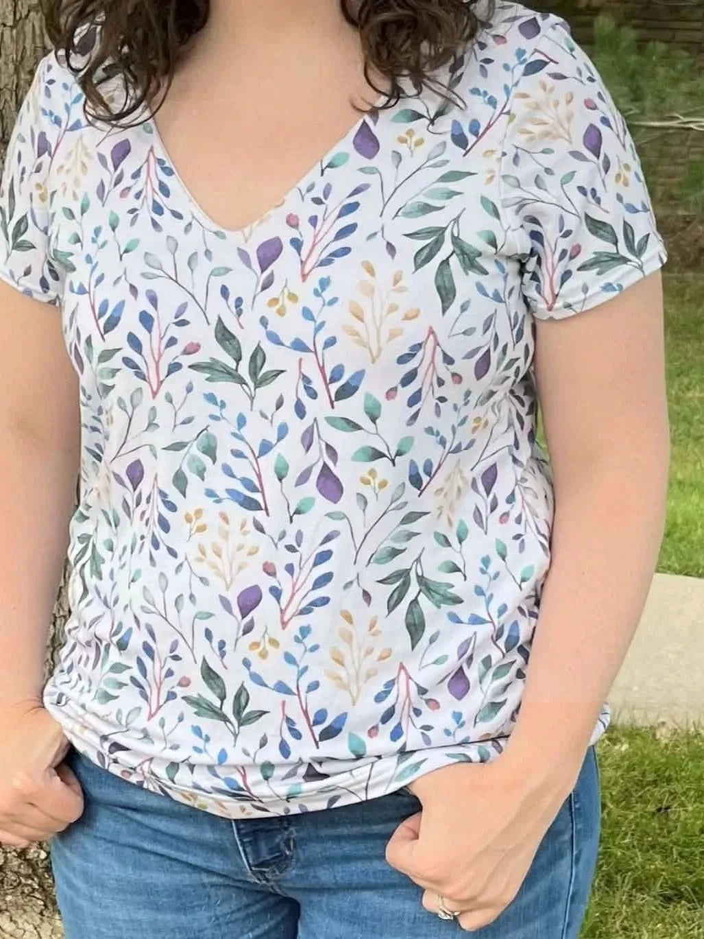 A person wears a white v-neck shirt with a colorful leaf pattern and blue jeans.
