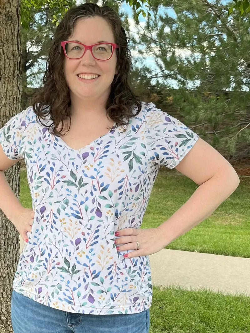 A woman with curly brown hair and red glasses smiles, wearing a white floral print shirt.
