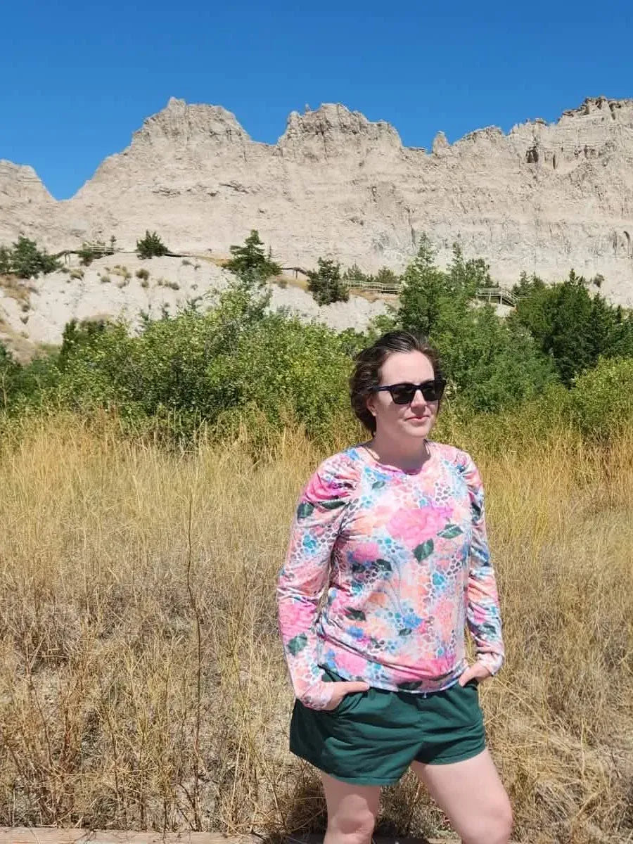 A woman in a floral long-sleeve shirt and dark shorts stands in a field.