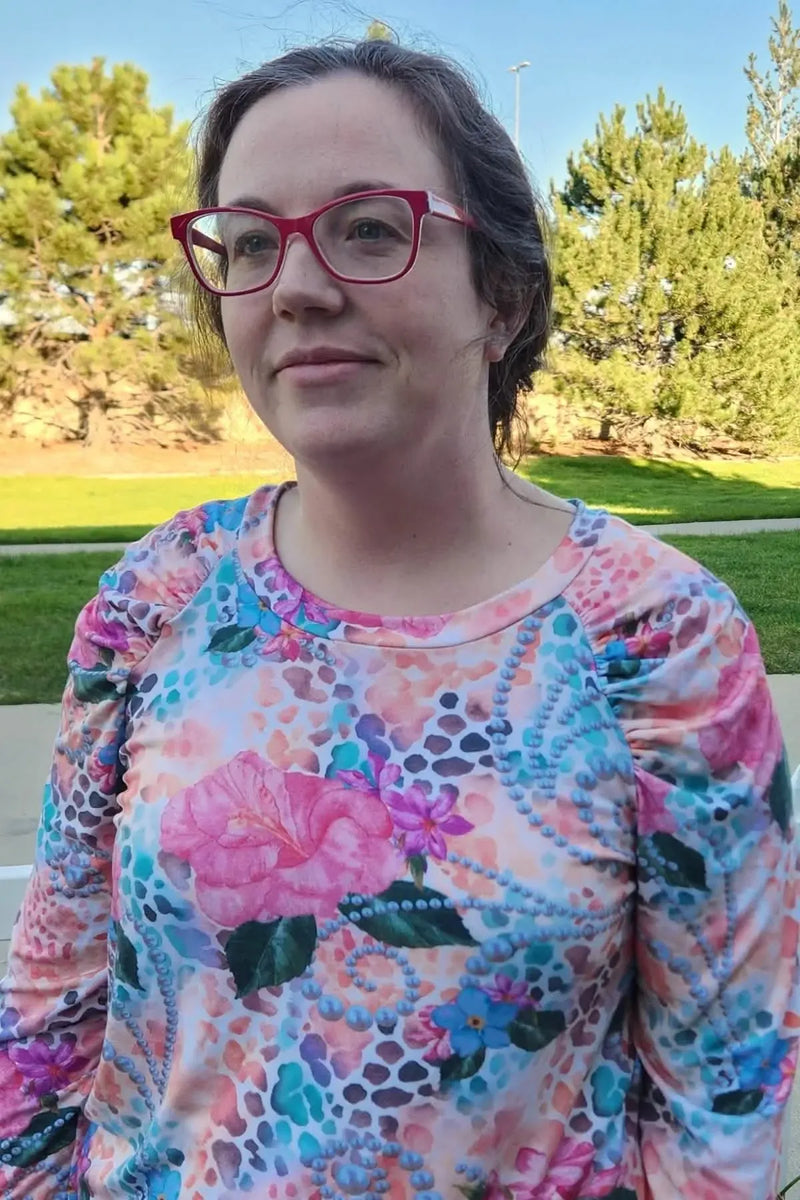 A woman wears a colorful floral and pearl print long-sleeved top with red glasses.