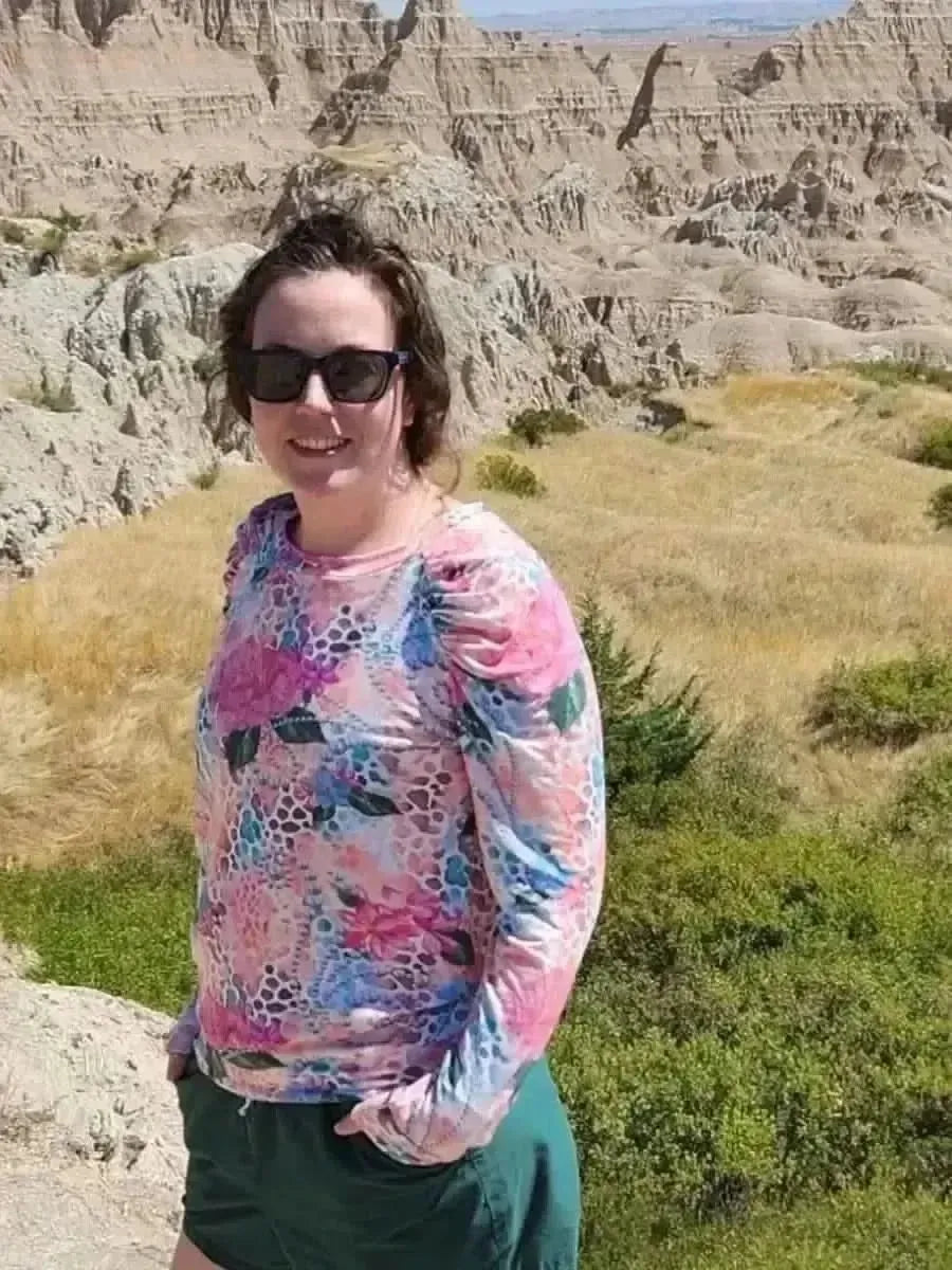Woman wearing a long-sleeved pink floral hibiscus print top with green shorts in a rocky landscape.