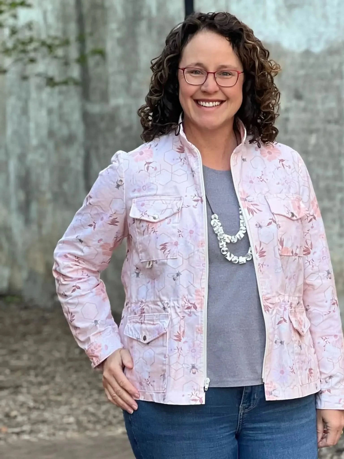 A woman smiles, wearing a peach floral jacket with bees, a gray shirt, and jeans.