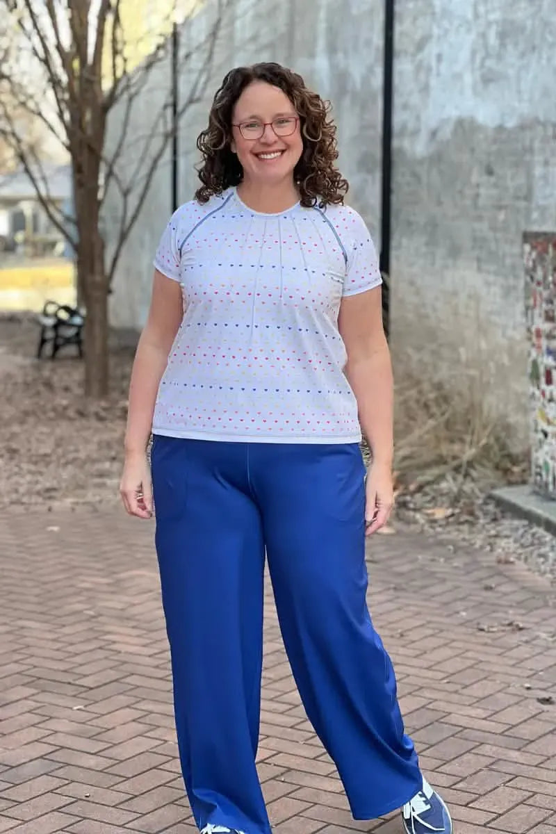 A woman smiles, wearing a white top with colorful hearts and blue pants.