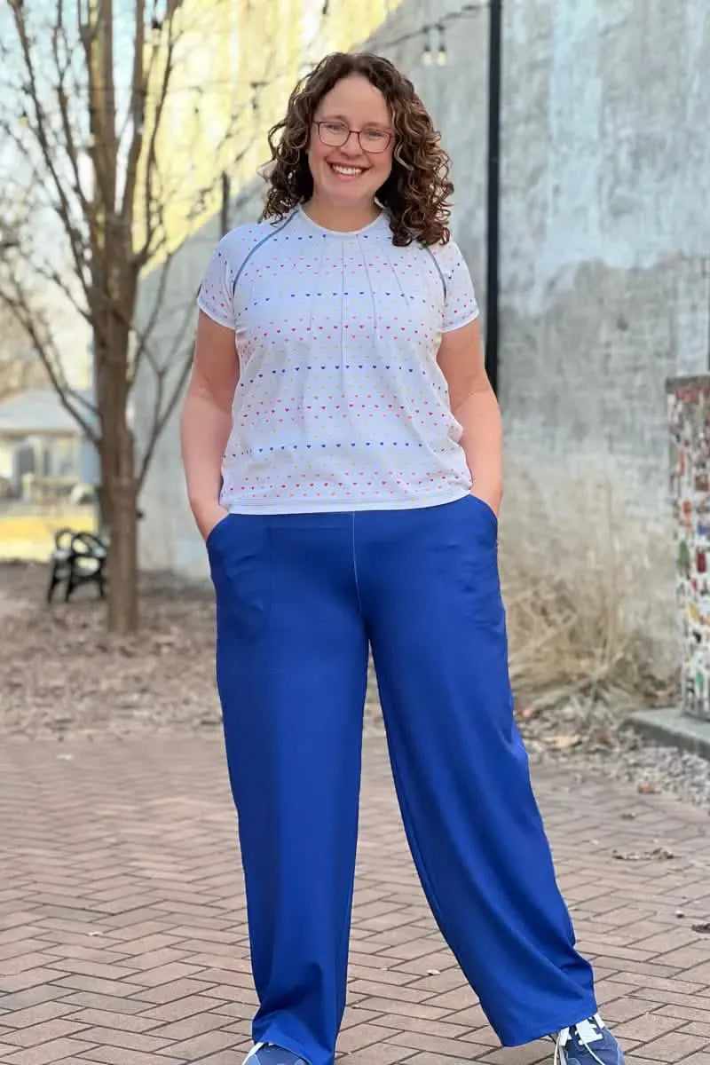 A woman smiles, wearing a white shirt with colorful hearts and blue pants.