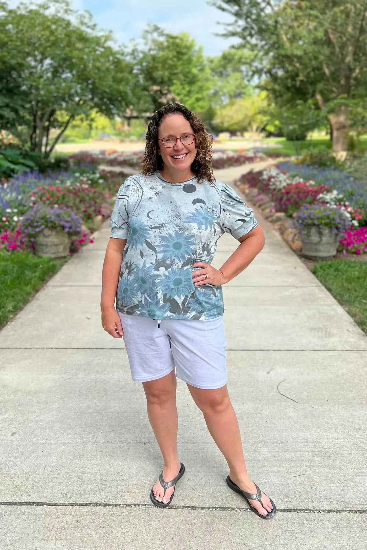 A woman smiles, wearing a light blue top with celestial sunflower print and white shorts.