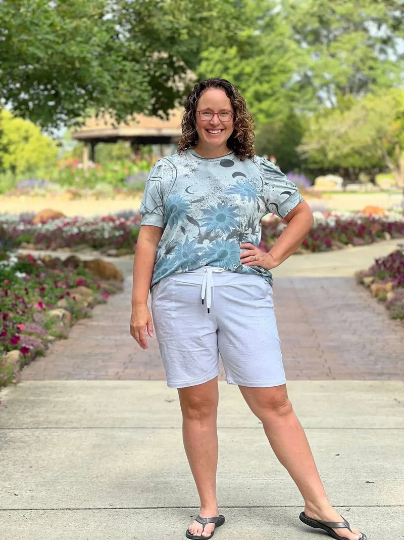 A woman in a blue floral top and light gray shorts stands on a paved path.