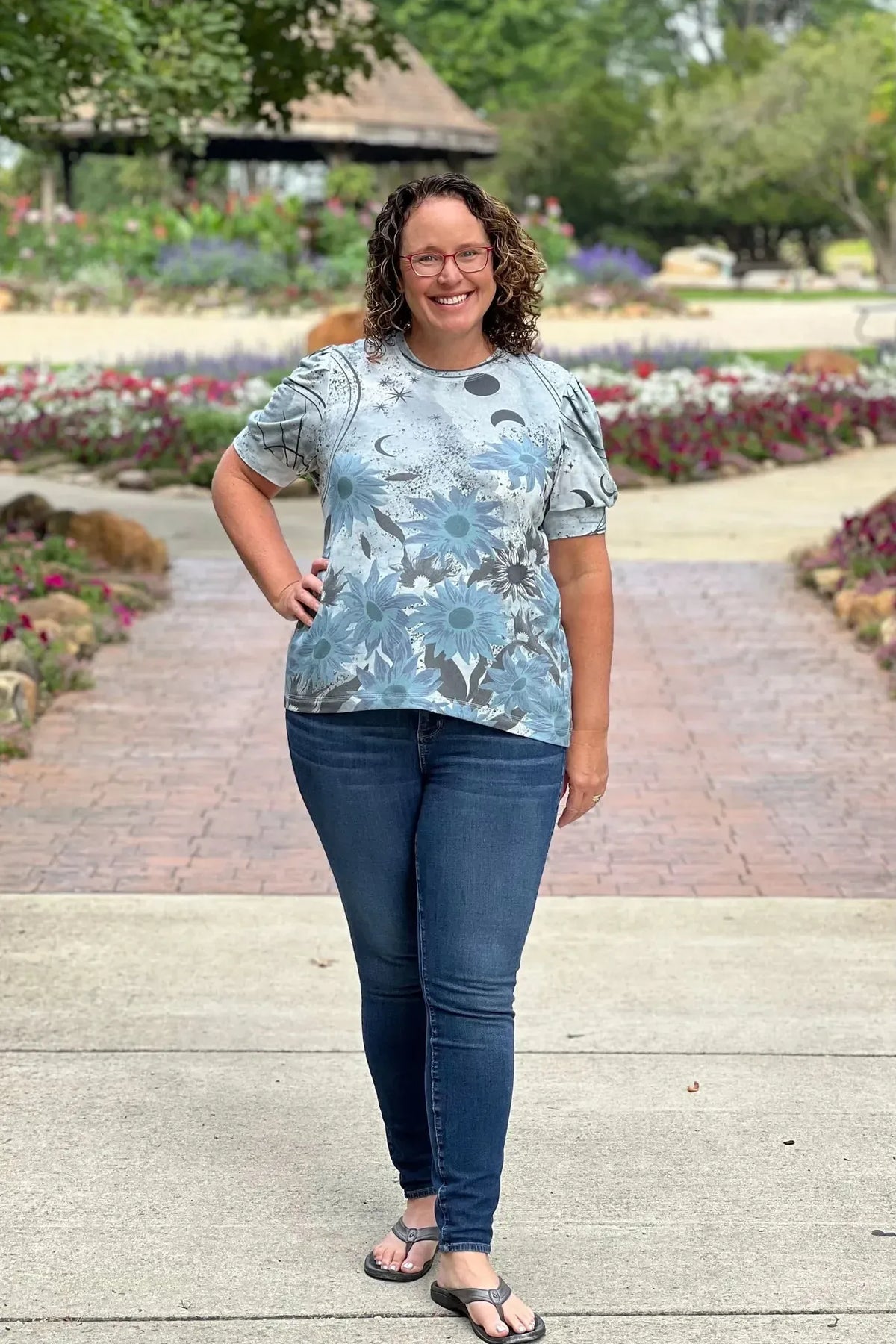 A woman wears a silver t-shirt with blue sunflowers and celestial designs.