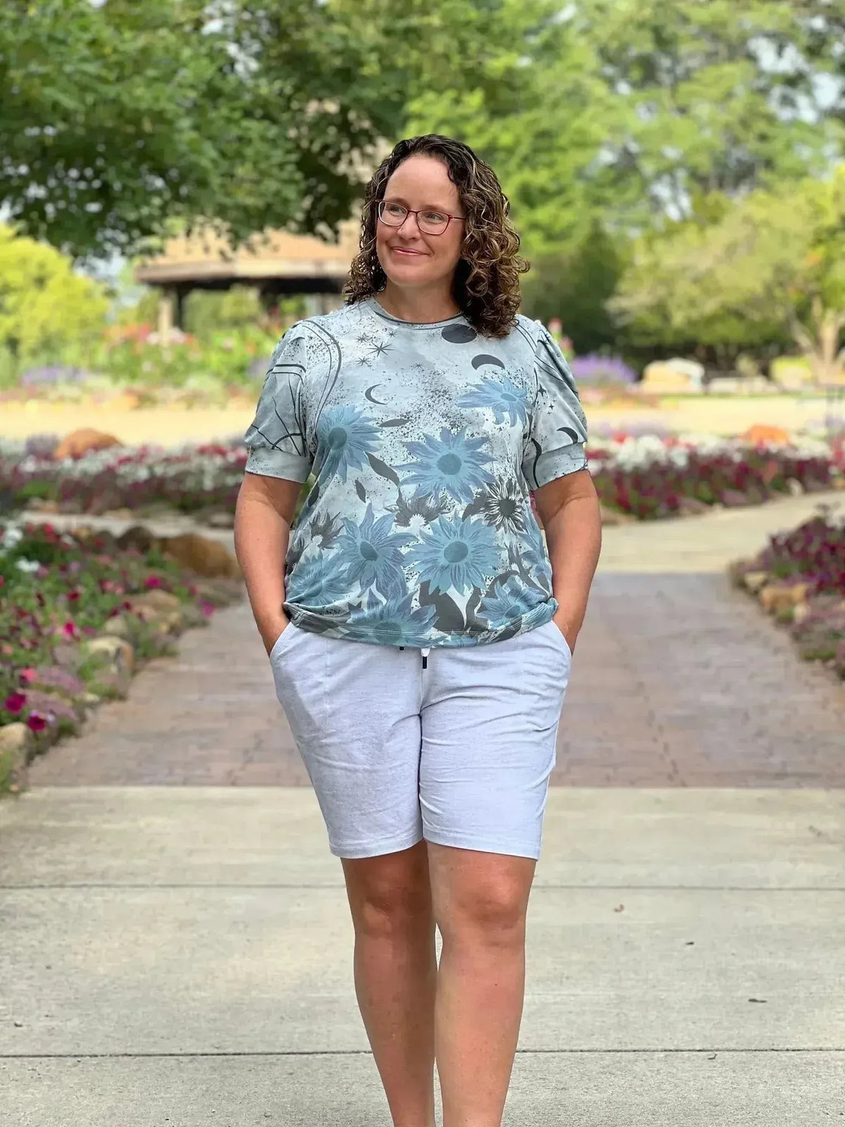 Woman modeling a blue Celestial Sunflowers Silver Panel top with linen shorts in a garden.