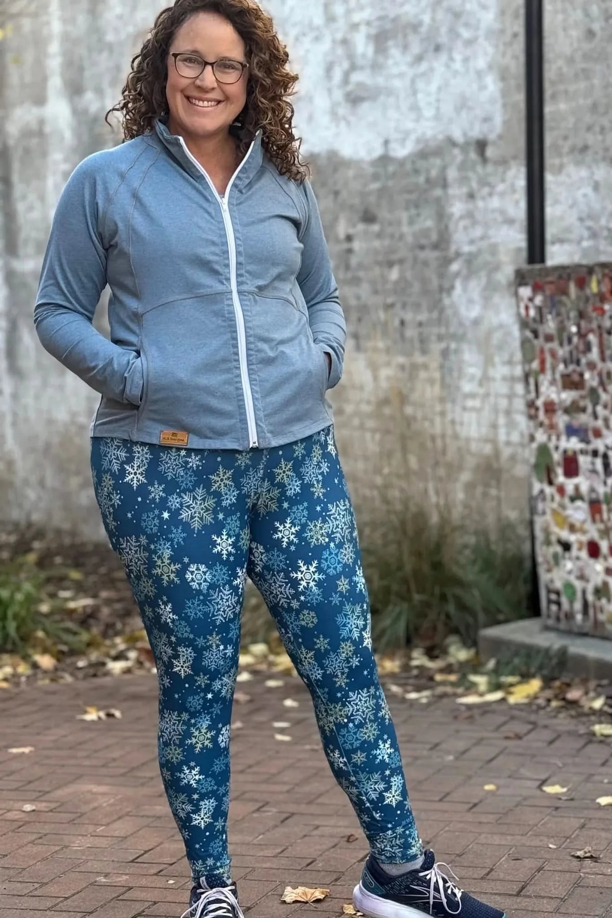 A woman smiles, wearing a gray zip-up jacket and blue snowflake-patterned leggings.