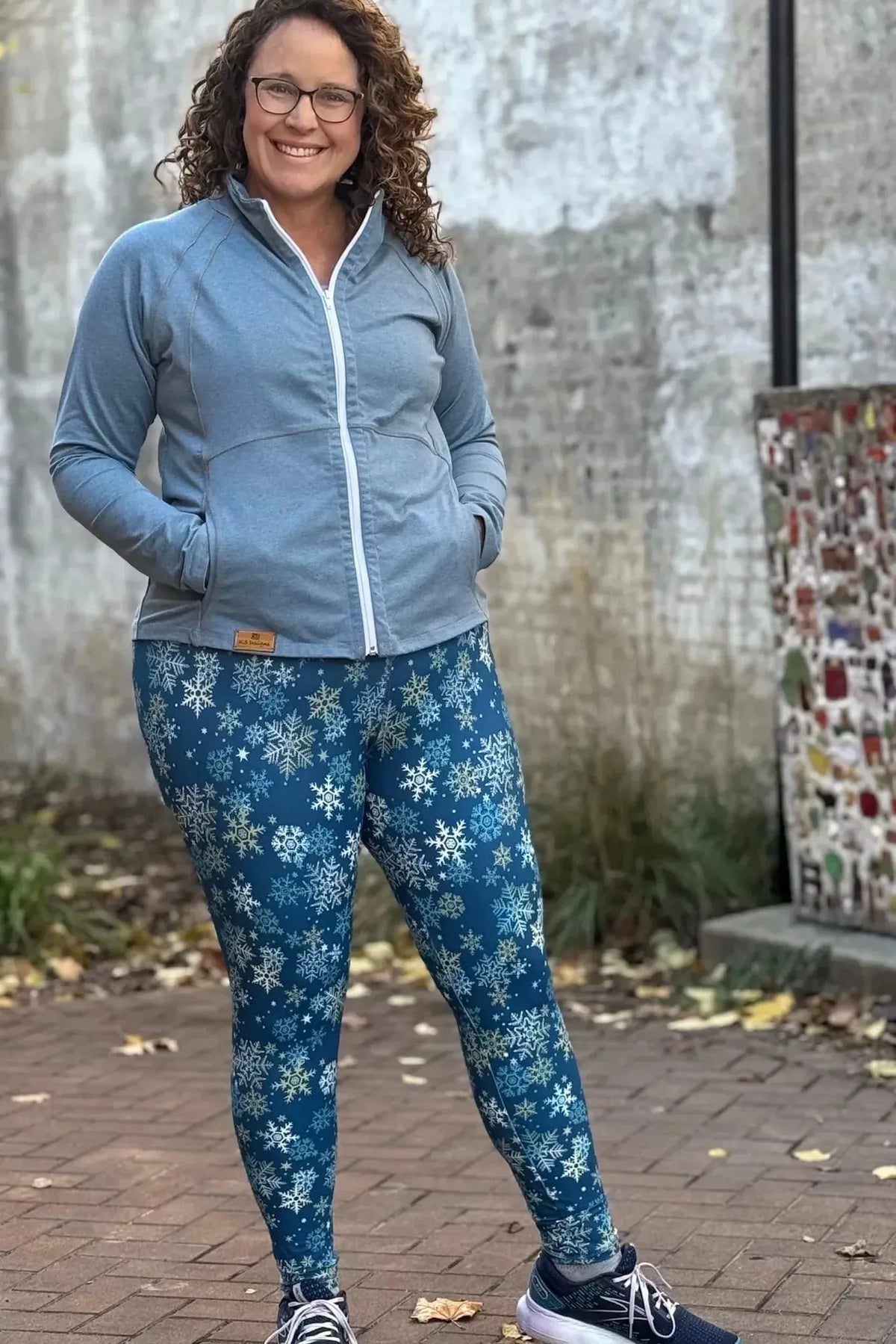 Woman modeling gray zip-up jacket and blue snowflake-print leggings outdoors.