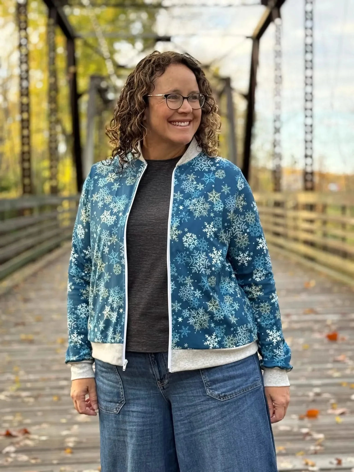 A woman smiles, wearing a blue snowflake-patterned bomber jacket and jeans.