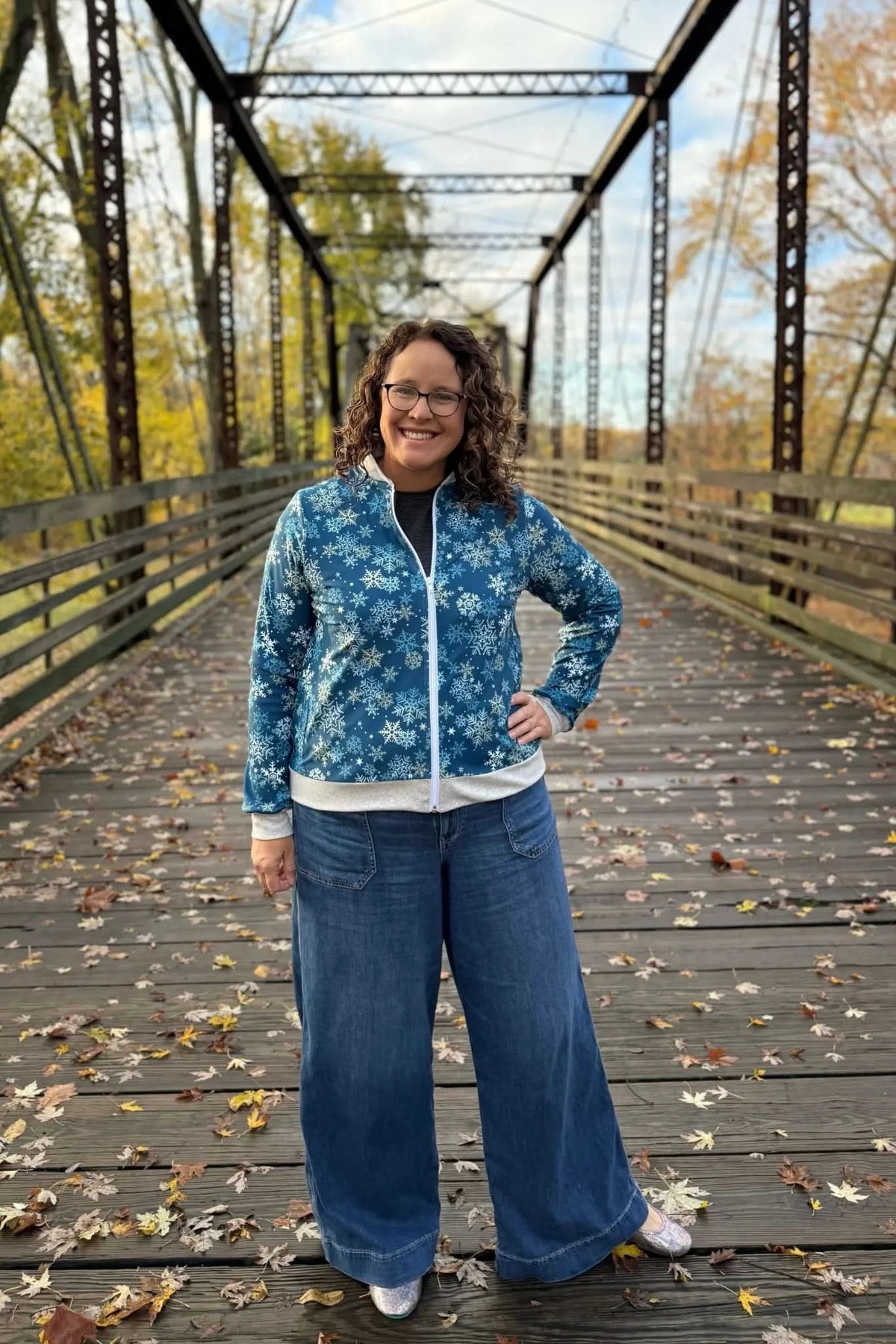 A woman smiles in a blue snowflake jacket and wide-leg jeans on a bridge.