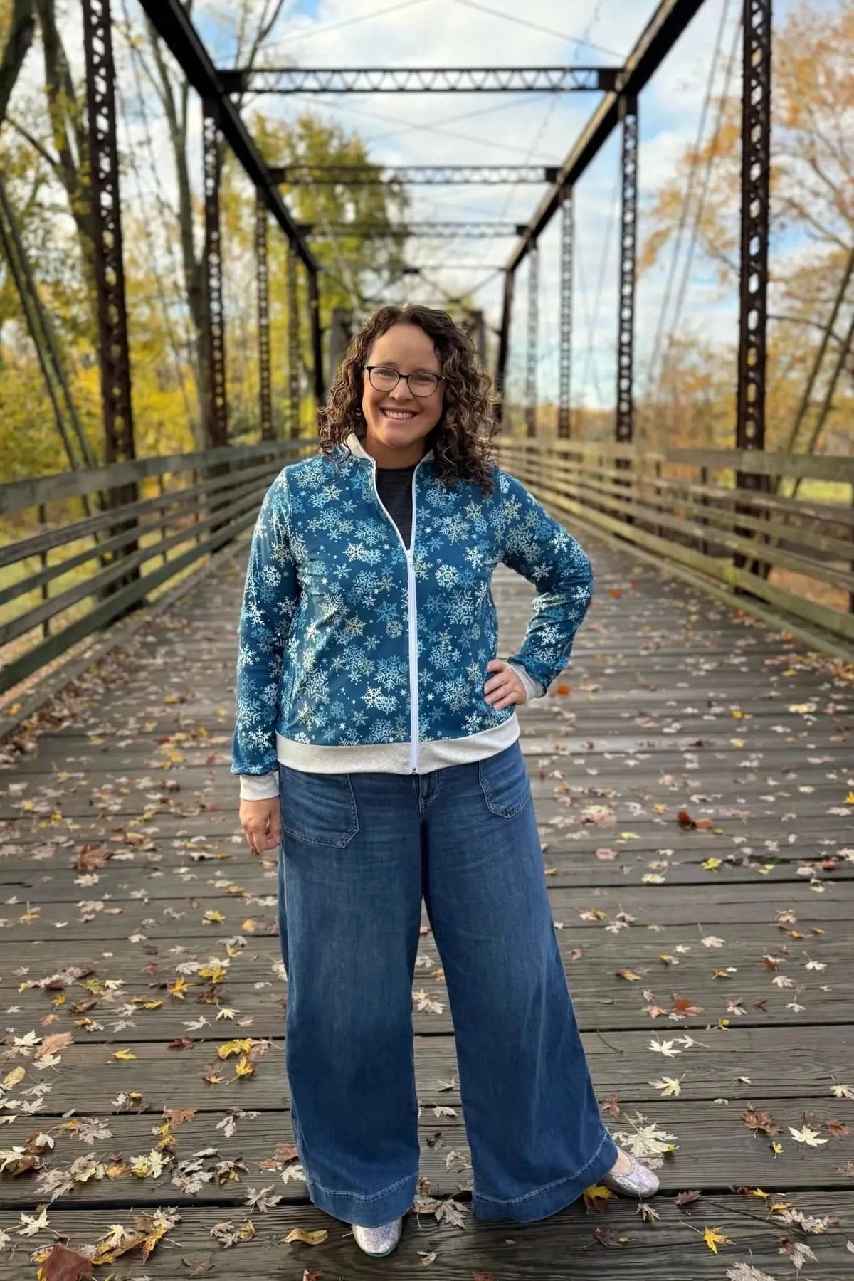 Woman wearing a blue snowflake-print zip-up jacket and wide-leg jeans on a wooden bridge.