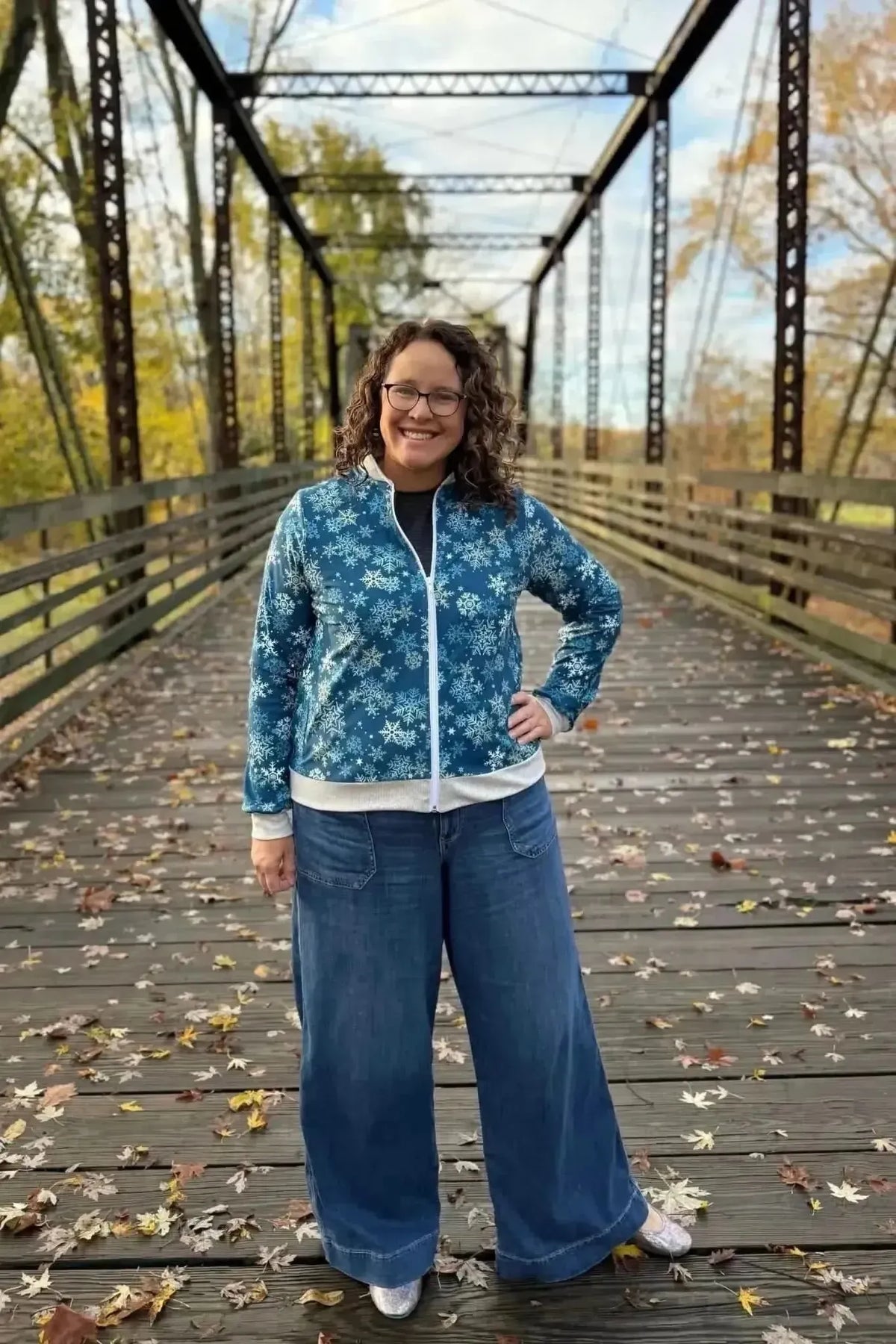 Woman wearing a blue snowflake-print zip-up jacket and wide-leg jeans on a wooden bridge.