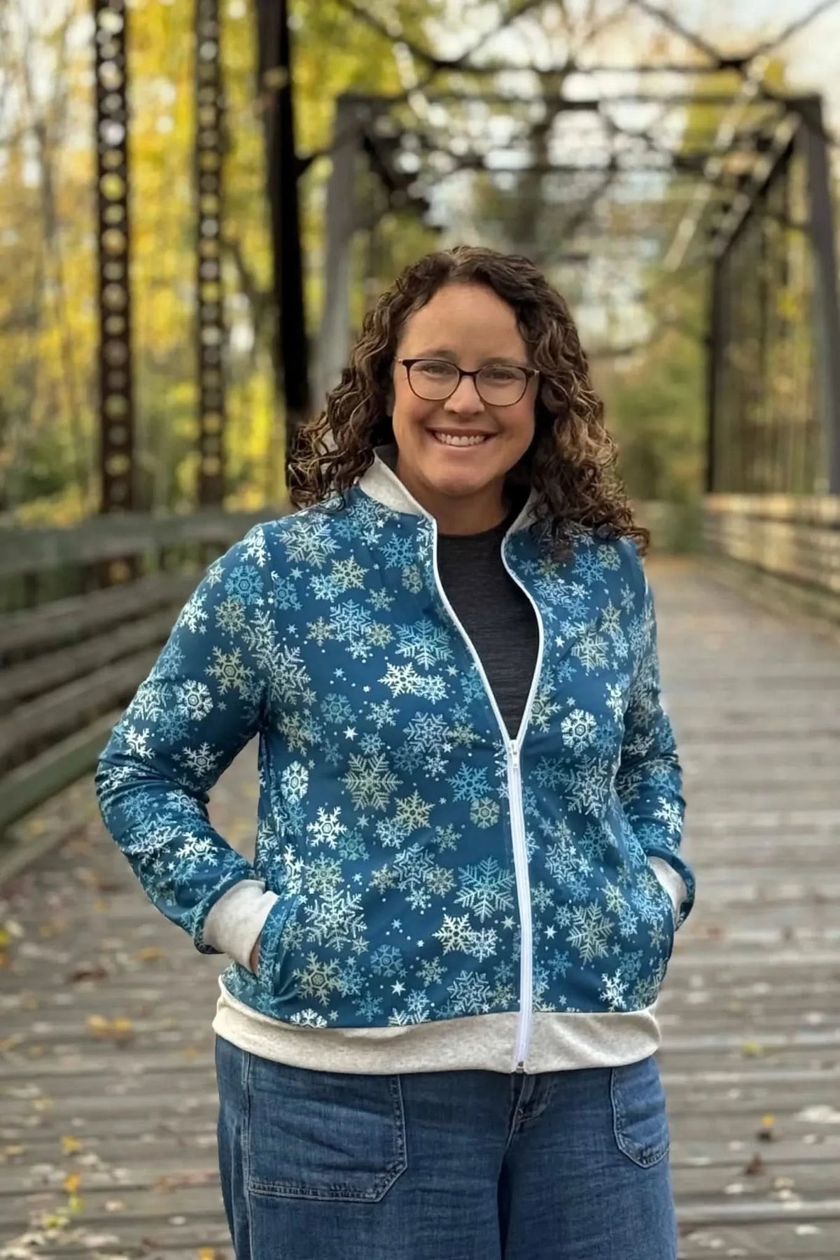 A woman smiles, wearing a blue snowflake-patterned jacket and jeans on a wooden bridge.