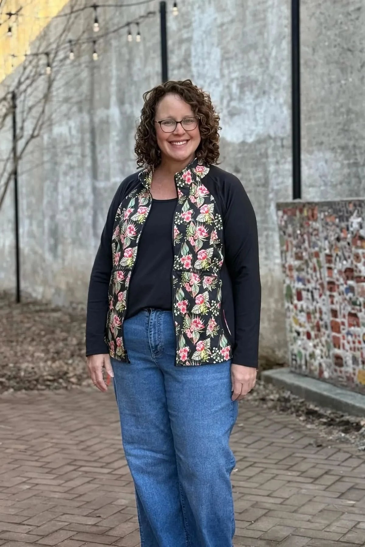A woman smiles, wearing a black long-sleeved shirt and a floral vest with pink and green flowers.