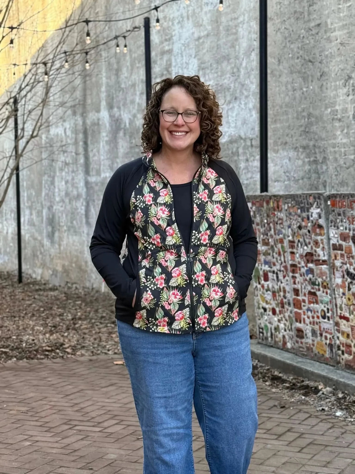 A woman in a black jacket with a floral print and blue jeans smiles at the camera.