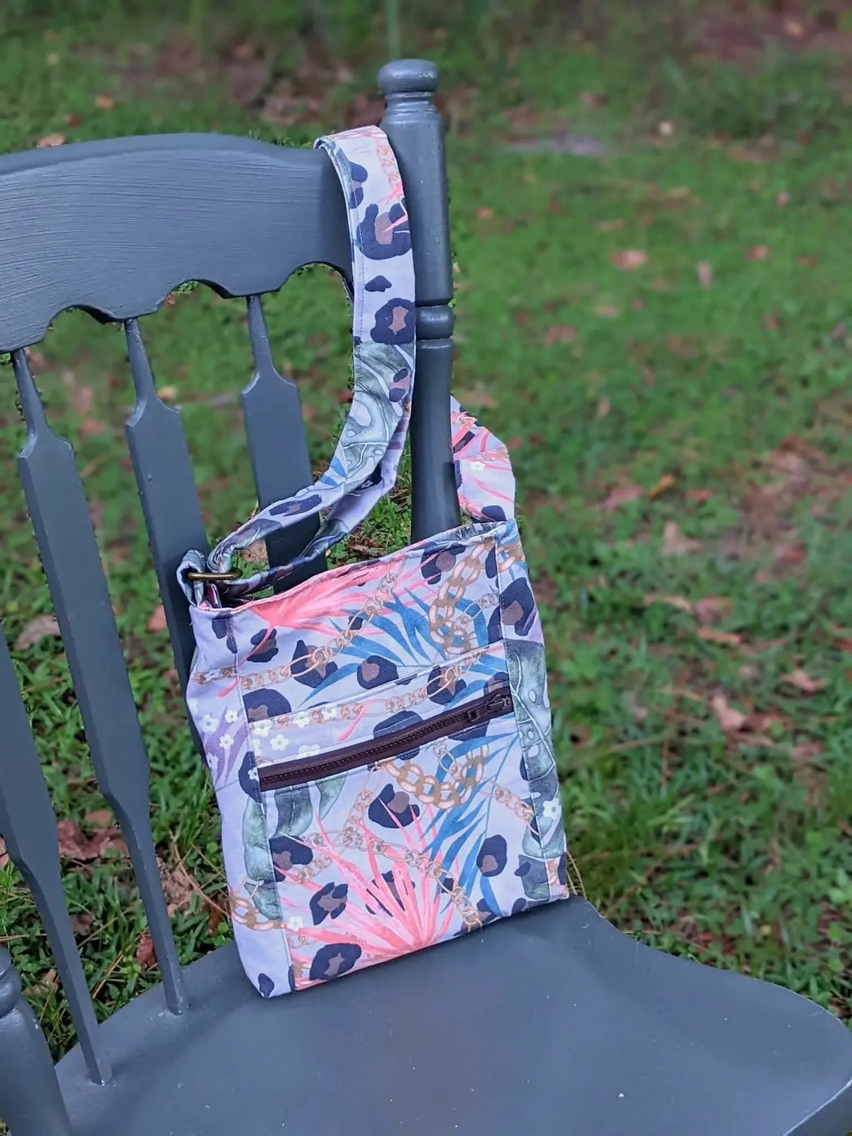 A crossbody bag with a leopard print and tropical leaf pattern hangs on a dark gray chair.