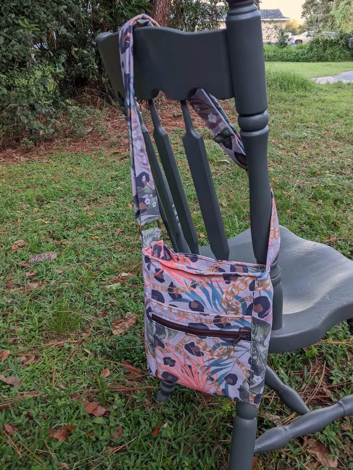 A crossbody bag with a leopard print and tropical leaf pattern hangs on a green chair.