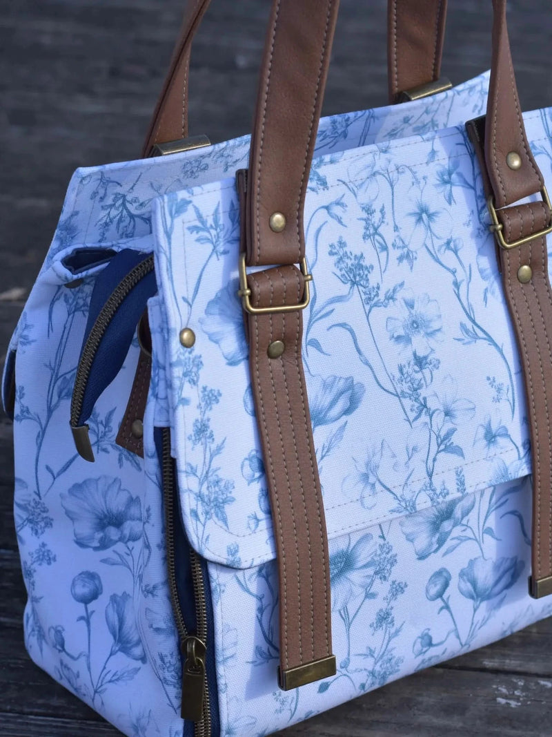 A blue and white floral patterned bag with brown leather straps and brass hardware.