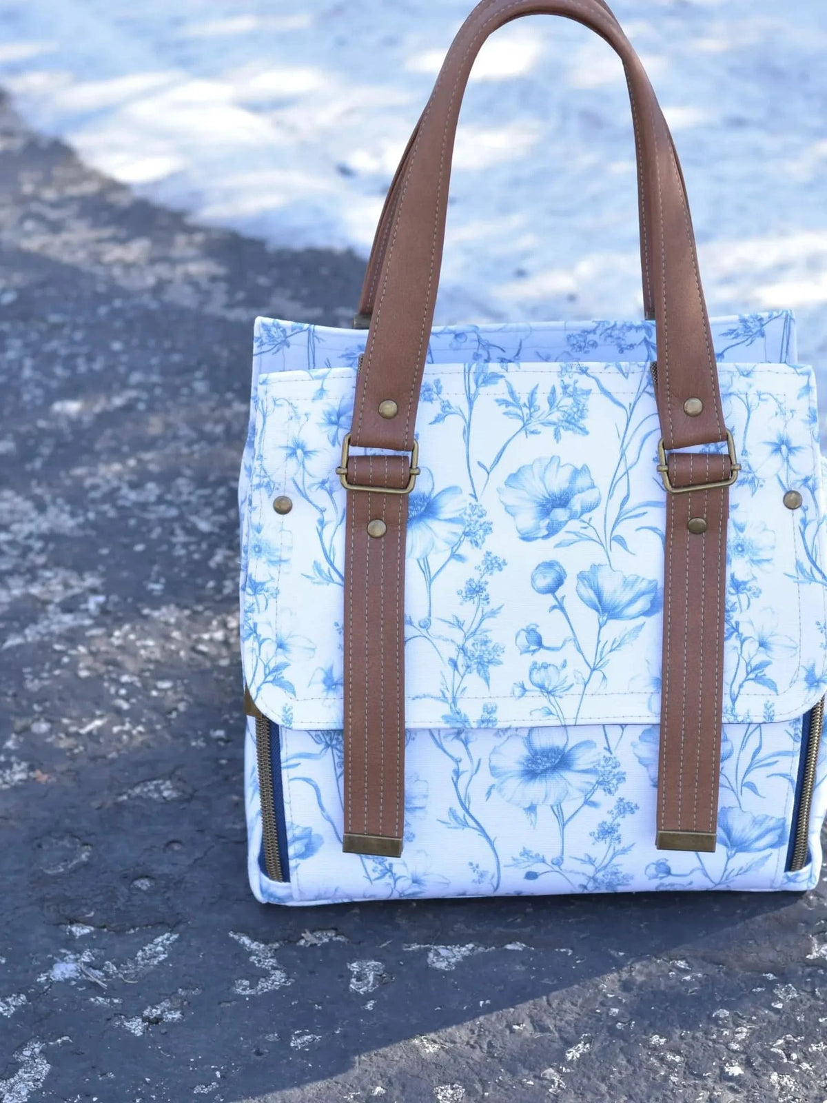 Blue and white floral bag with brown straps on a snowy ground.