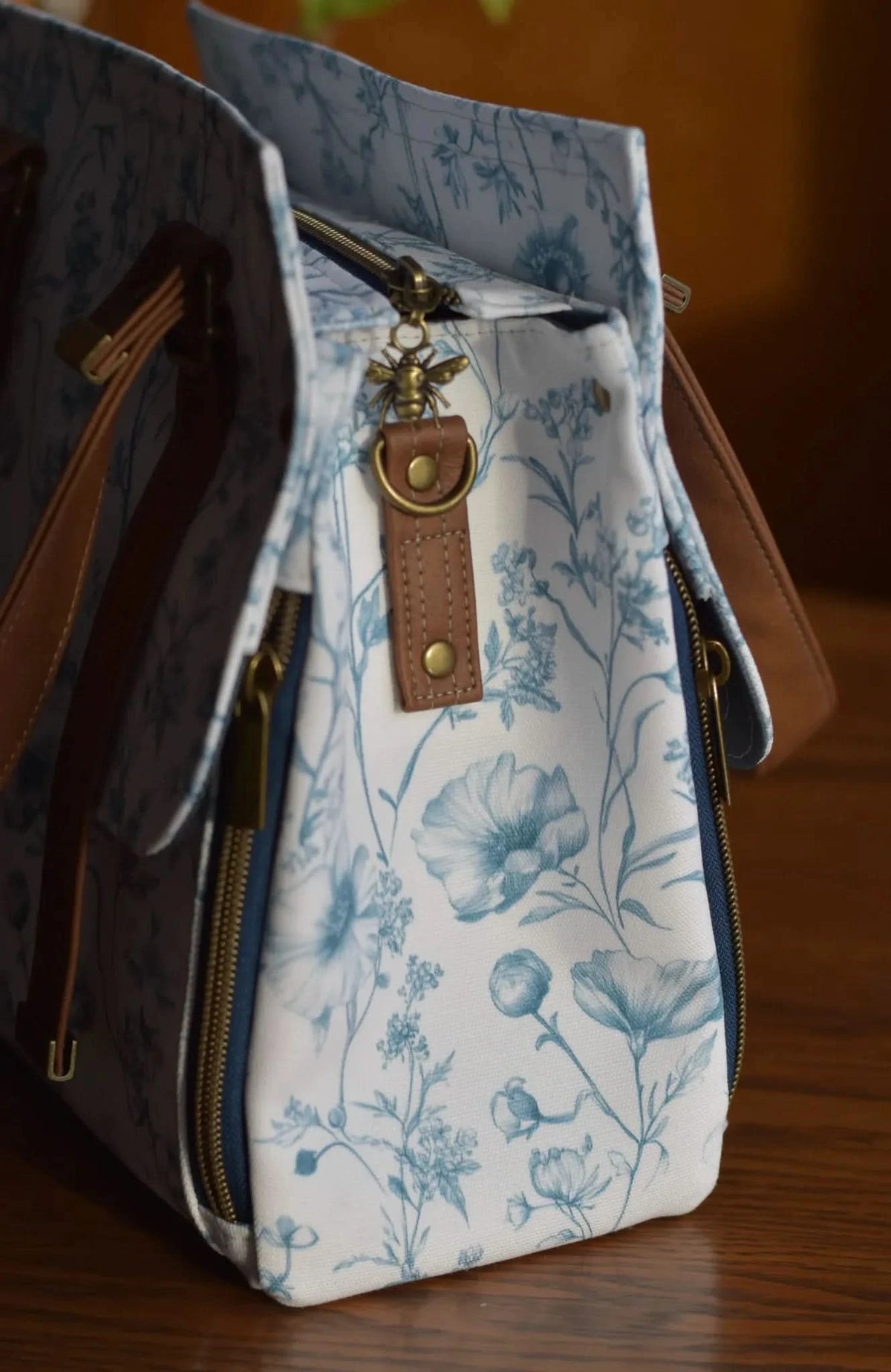 A white bag with blue floral patterns, brown straps, and a bee charm.