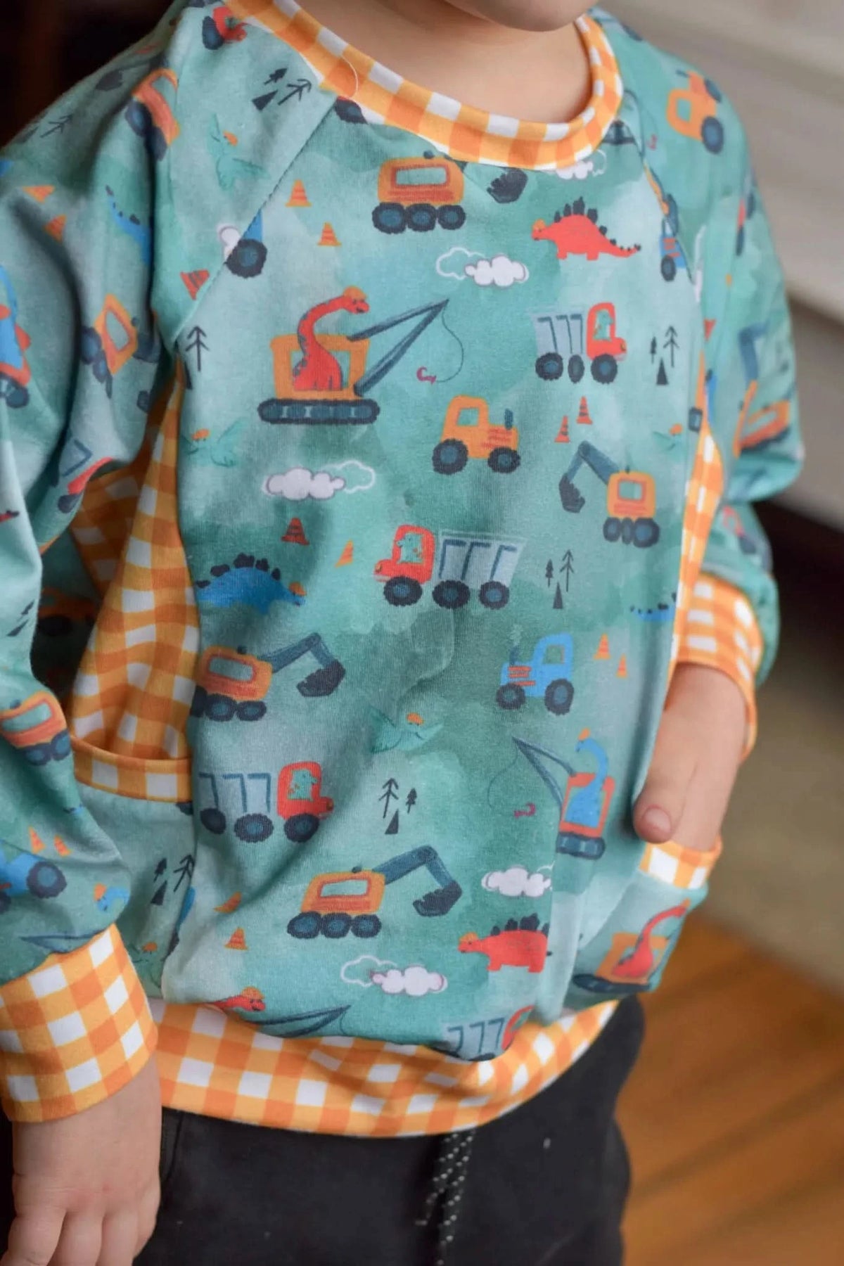 A child wears a green shirt with construction vehicles and dinosaurs, and orange gingham trim.
