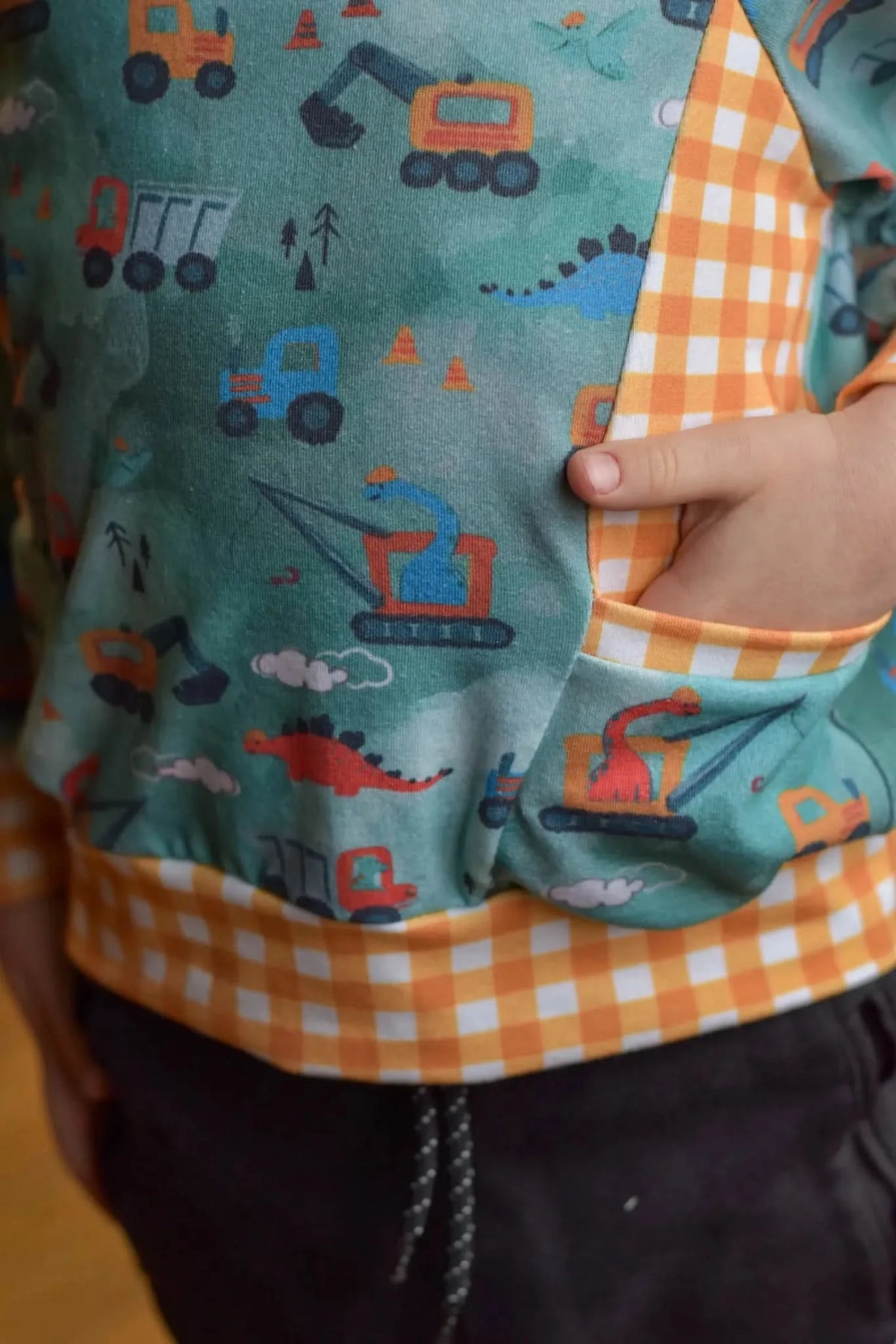A child wears a green shirt with construction vehicles and dinosaurs, with a checkered pocket.