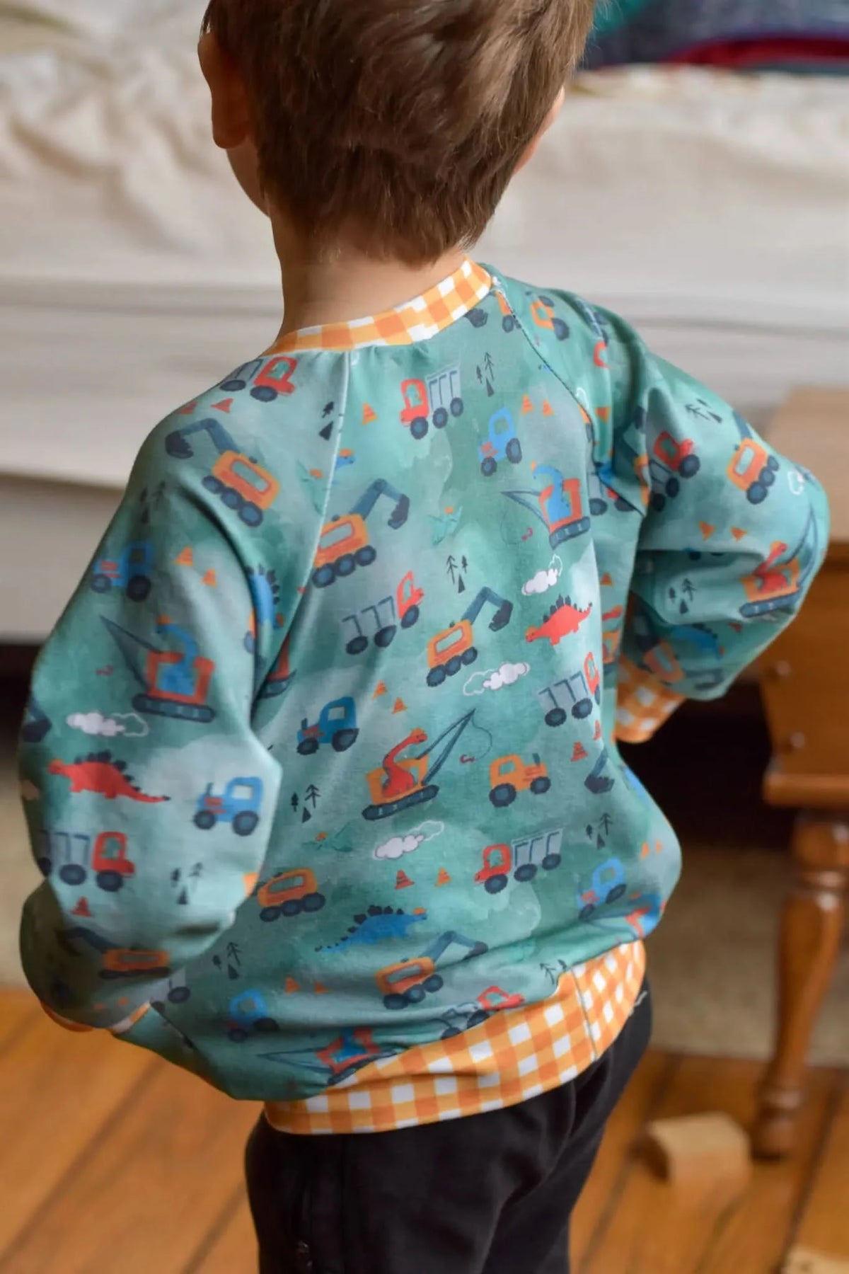 A child from behind wearing a green shirt with construction vehicles and dinosaurs.