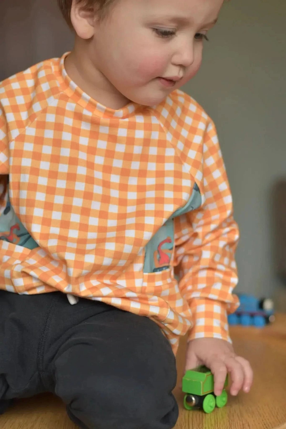 Child in a mustard gingham shirt with a dinosaur pocket, playing with a green toy train.