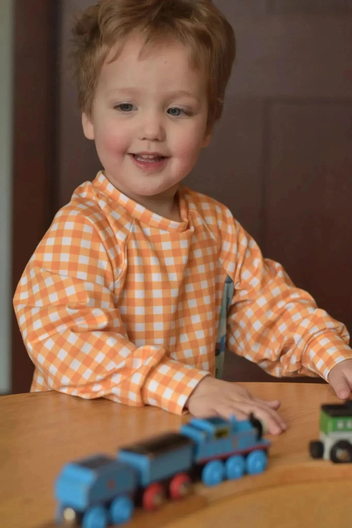 A child in a mustard gingham shirt plays with toy trains.