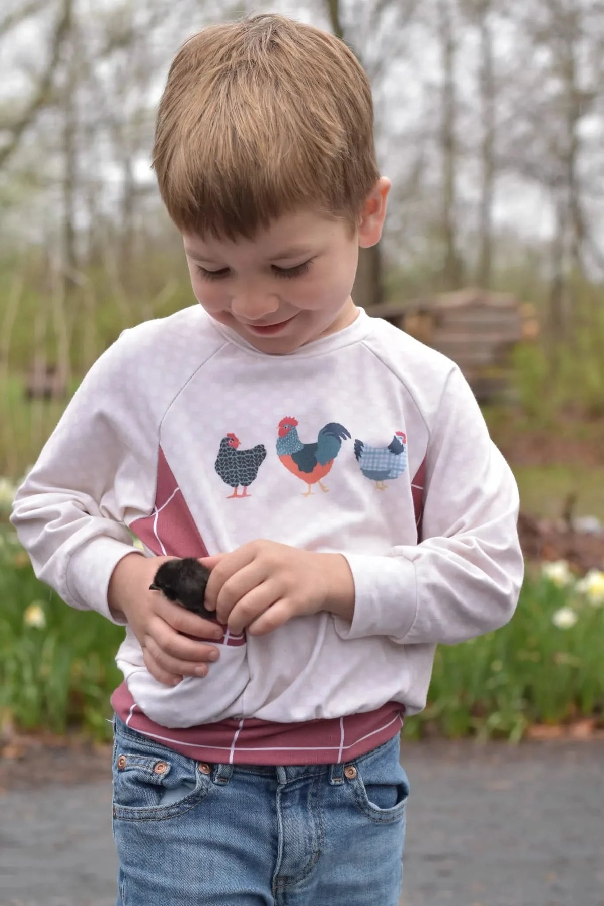 A child in a white shirt with patchwork chickens holds a small black chick.