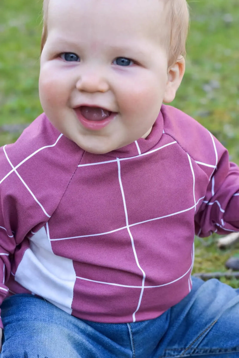 A baby wears a red windowpane shirt with white lines and blue jeans.