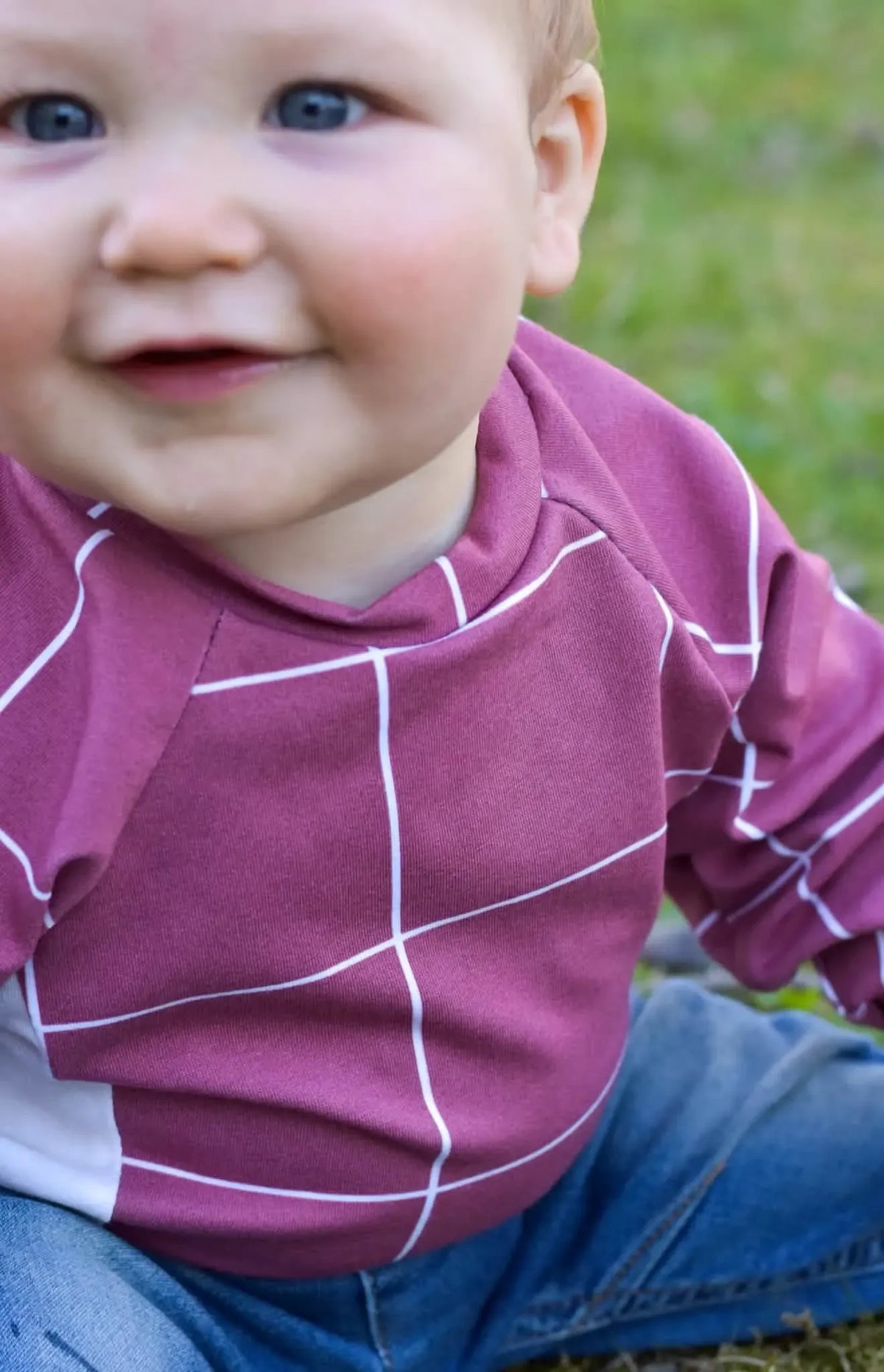 A baby wears a red windowpane shirt and blue jeans, smiling at the camera.