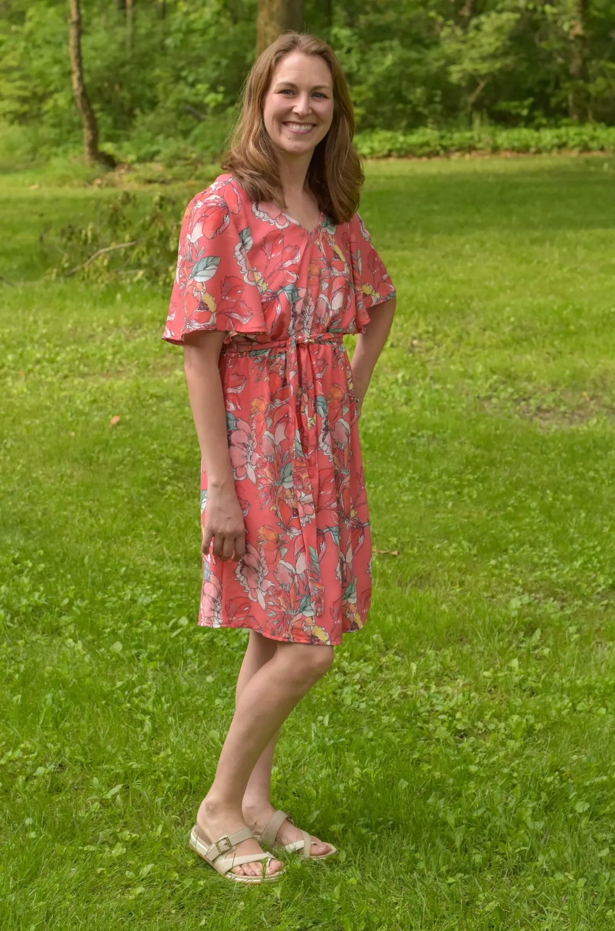 A woman in a coral floral dress with a tie waist stands in a grassy field.