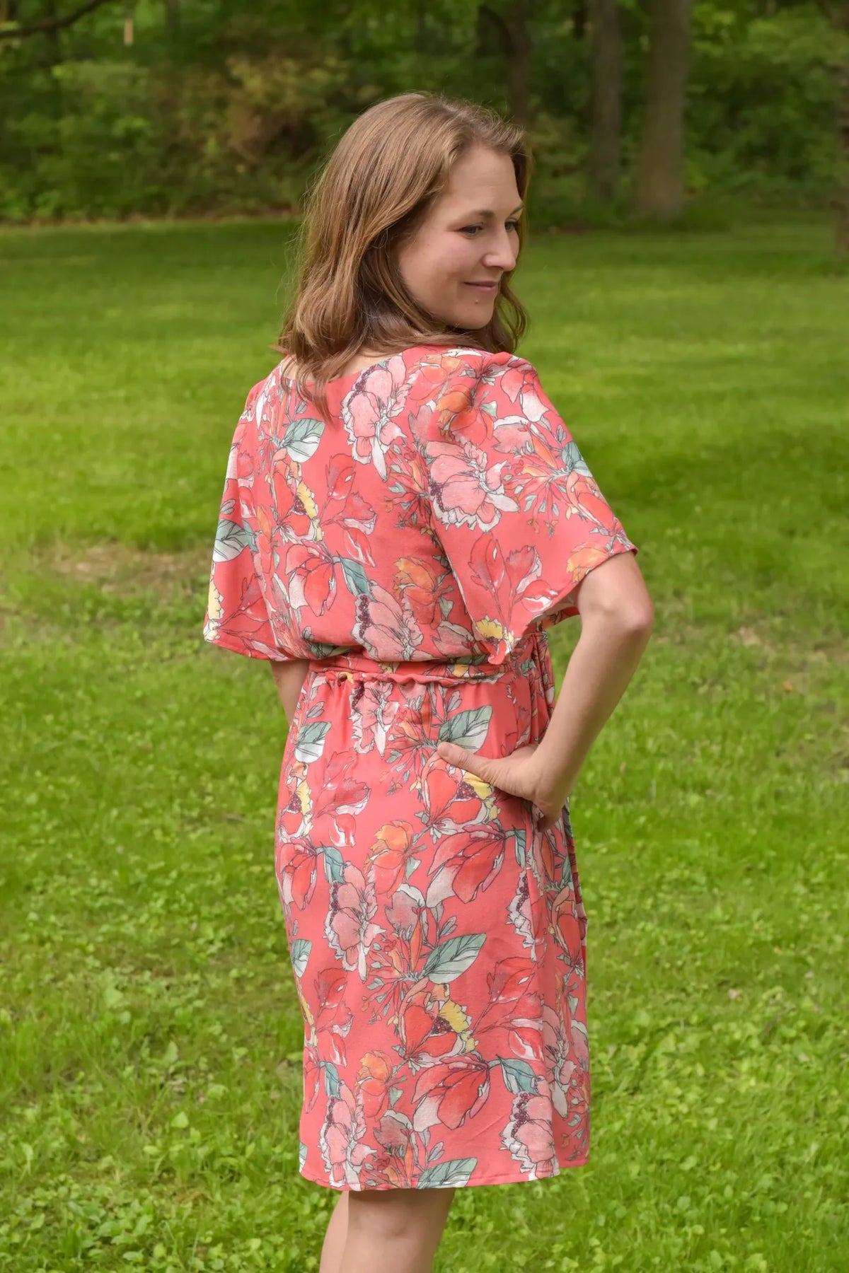 Woman in a coral floral dress with her hand on her hip, looking over her shoulder.